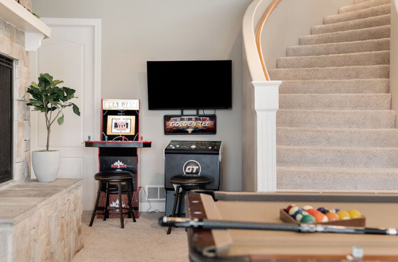 Een biljarttafel met een keu, een aan de muur bevestigde televisie, een bordje voor een Golden Tee spel en een trap.