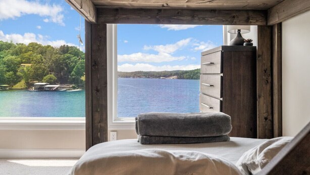 Een slaapkamer met een groot raam met uitzicht op een meer en bos. Er staat een bed met een grijs kussen en een houten dressoir met een lamp.