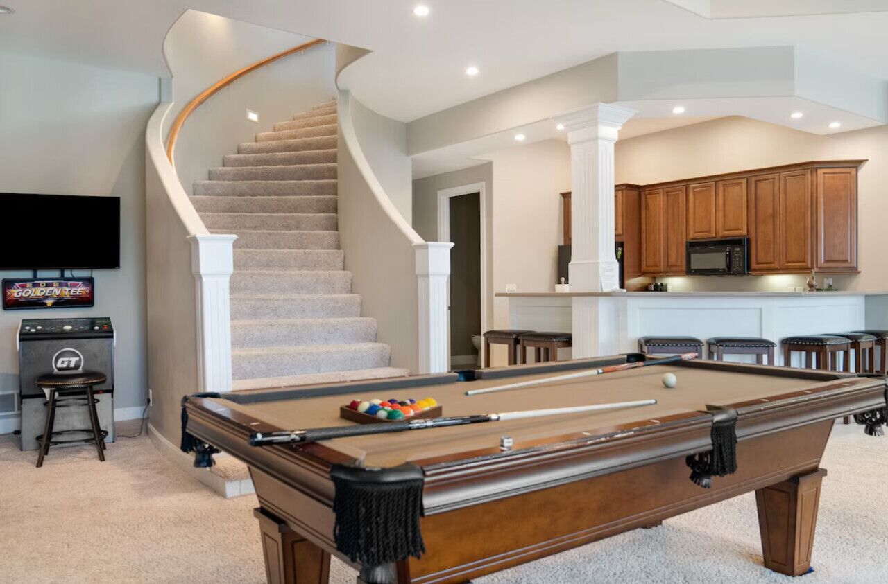 Une table de billard dans une pièce dotée d’un escalier à volutes, de meubles en bois et d’un téléviseur à écran plat.