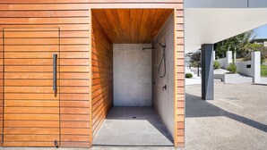 Ducha exterior moderna con paneles de madera y suelo de baldosas, ubicada en un patio con un edificio al fondo.