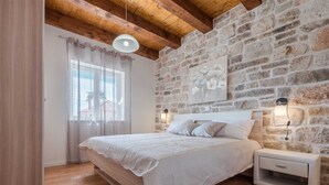 A bedroom with a stone wall, a bed with a white bedspread, a nightstand, and a window with sheer curtains. - Stone Villa Gelosia Rustica with heated pool (Sukošan)