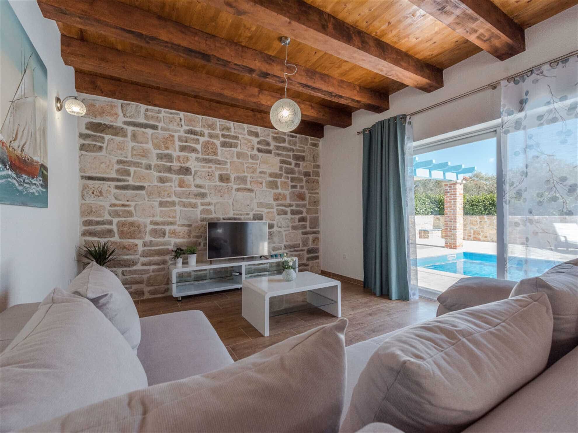 Ein modernes Wohnzimmer mit Steinwand, Holzbalkendecke, Flachbildfernseher und Blick auf einen Pool.