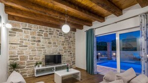 A living room with a stone wall, wooden ceiling beams, a TV, and a sofa. - Stone Villa Gelosia Rustica with heated pool (Sukošan)