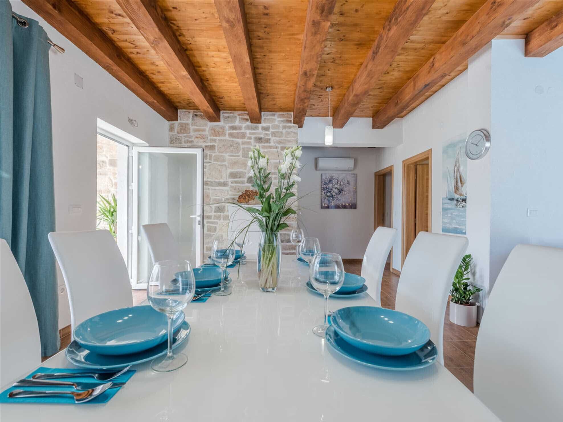 Une salle à manger avec un mur en pierre, des poutres en bois au plafond et une table dressée pour six personnes, avec des assiettes bleues et des verres à vin.