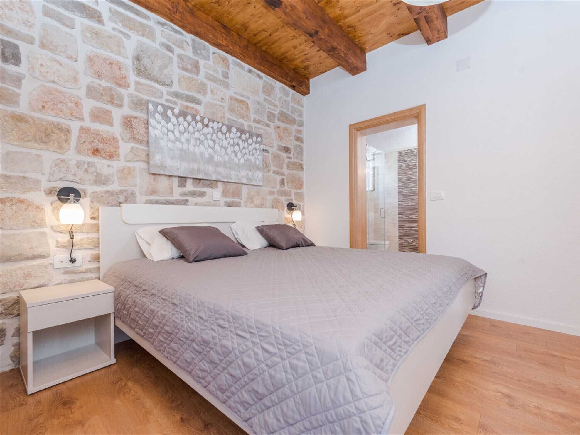 A bedroom with a stone wall, wooden ceiling beams, a bed with a quilted cover, two bedside lamps, and a nightstand.