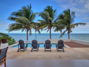 A poolside area with four Adirondack chairs facing a pool, surrounded by palm trees and overlooking the ocean. - Casa Midtown  -  Yucatan Home Rentals (Chicxulub Puerto)