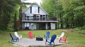 Une maison à deux étages avec un balcon, entourée d’une forêt. On y trouve six chaises Adirondack disposées en cercle sur une surface recouverte de gravier, devant la maison.