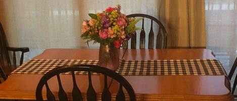A wooden dining table with a checkered placemat, surrounded by four chairs with a unique back design. A vase with flowers is placed on the table.