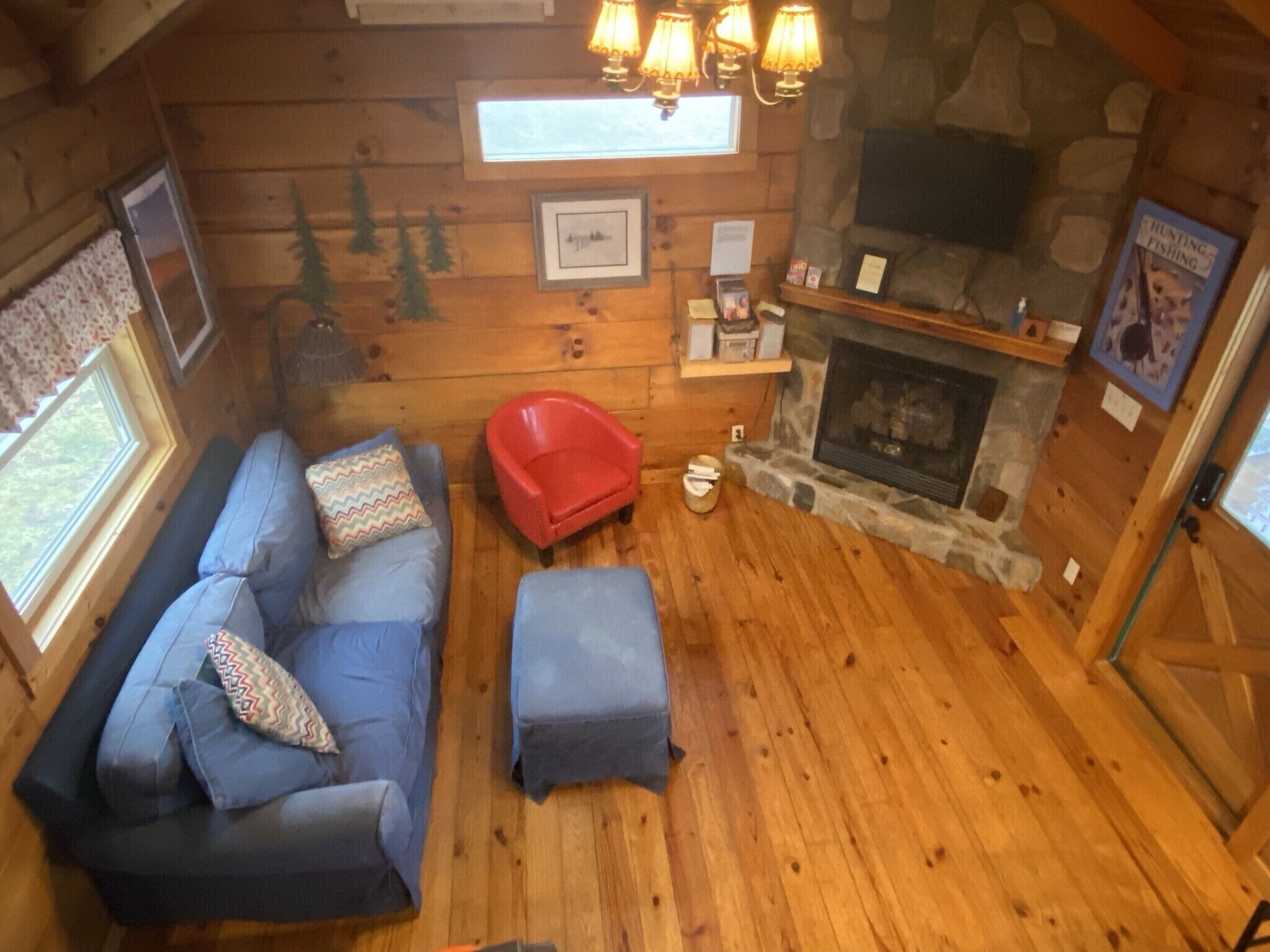 A cozy cabin interior with a wooden floor, a blue sofa set, a red chair, a fireplace, and a stone wall.