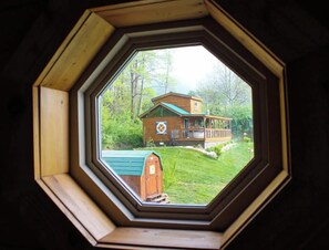 Vista através de uma janela octogonal de uma casa com telhado verde, um deck de madeira e um gramado bem cuidado.