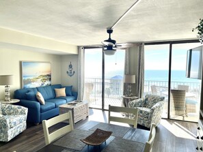A living room with a blue sofa, a white chair, a small table, and a view of the ocean through large windows. - GS Surf and Racquet 707A   Large Remodeled 2 bd / 2 ba  south facing (Gulf Shores)