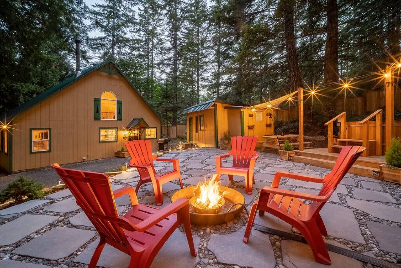 A cozy outdoor seating area with red chairs around a central fire pit, set against a backdrop of wooden cabins and a forested landscape.