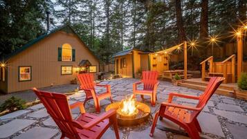 A cozy outdoor seating area with red chairs around a central fire pit, set against a backdrop of wooden cabins and a forested landscape.