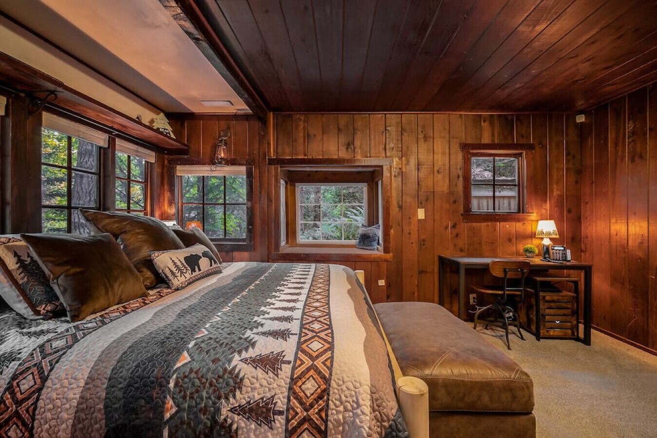 A cozy log cabin bedroom with a large bed, a desk, and a window overlooking the forest.