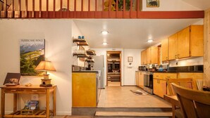 Una cocina compacta con armarios de madera, un comedor con mesa y sillas, y una escalera que lleva a un nivel superior.