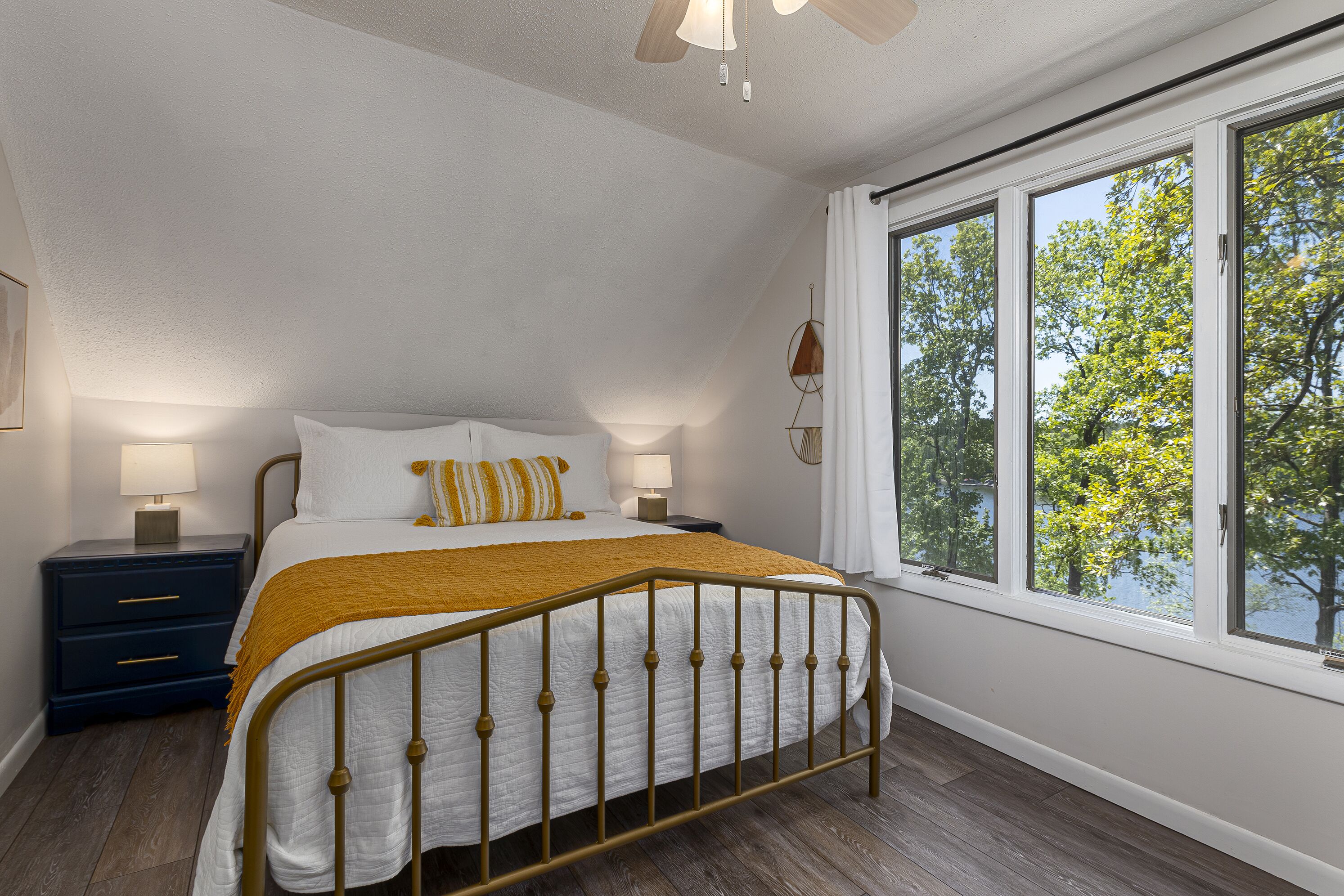 Een slaapkamer met een bed, nachtkastjes, een raam met uitzicht op bomen en een plafondventilator.