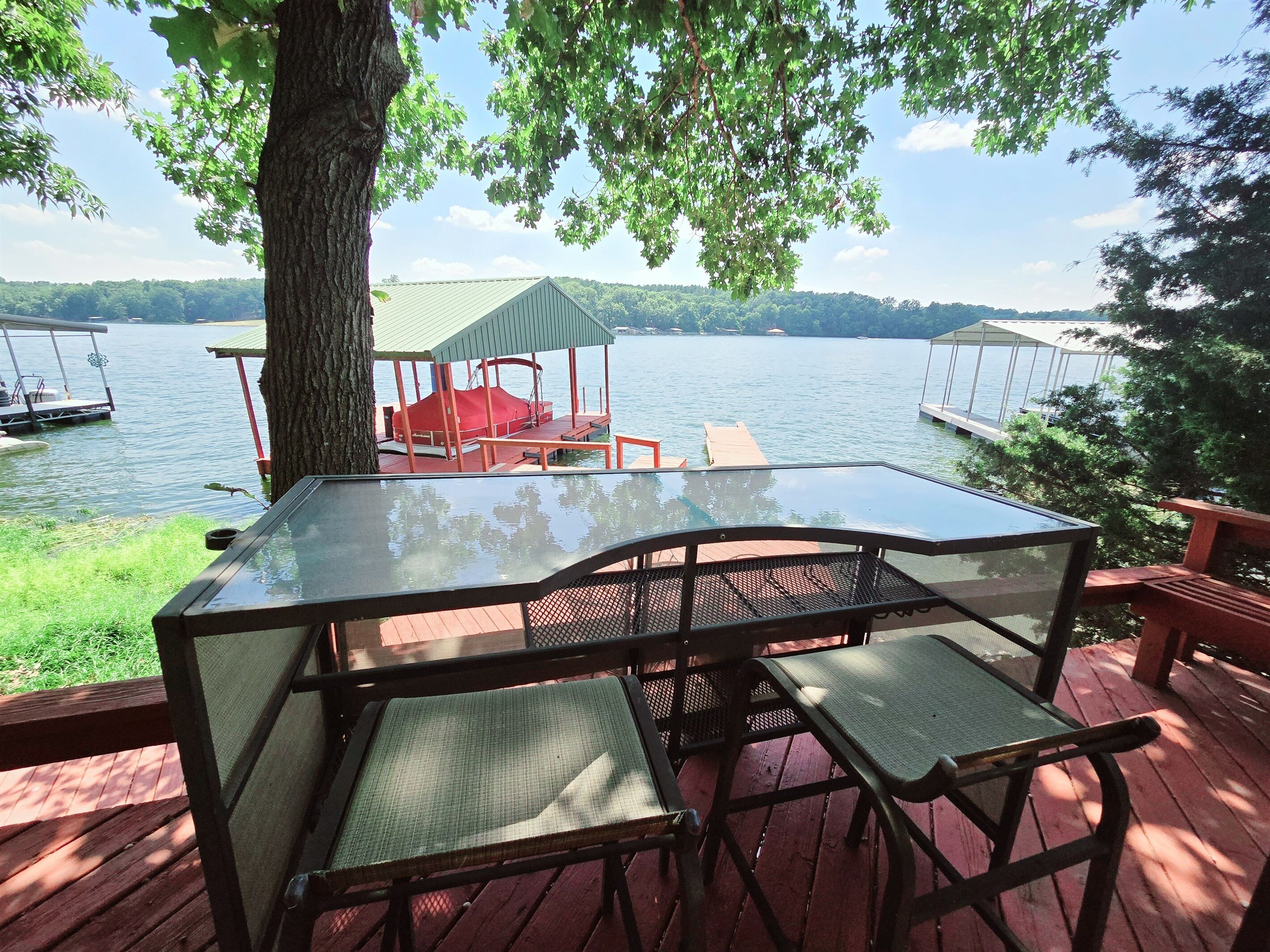 A glass-top table with two chairs on a wooden deck overlooking a lake with docked boats and a covered seating area.
