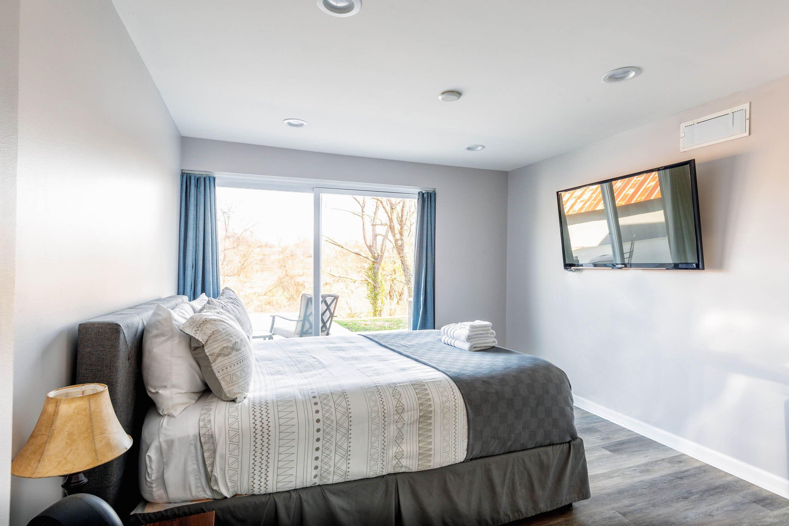 A modern bedroom with a large bed, a bedside table with a lamp, a flat-screen TV mounted on the wall, and a window with curtains.