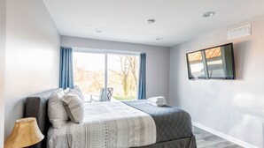 A modern bedroom with a large bed, a bedside table with a lamp, a flat-screen TV mounted on the wall, and a window with curtains.