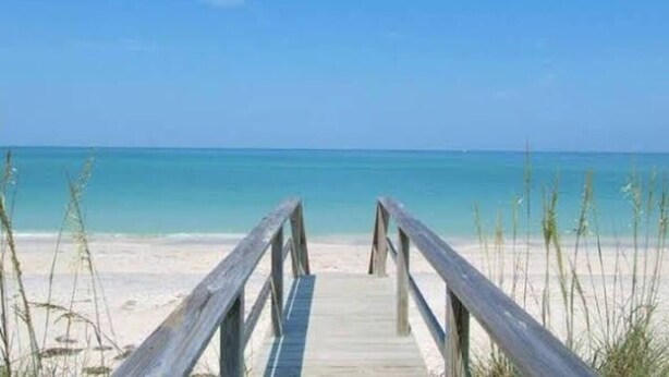 Ein hölzerner Steg führt zu einem Strand mit klarem, blauem Wasser und blauem Himmel.