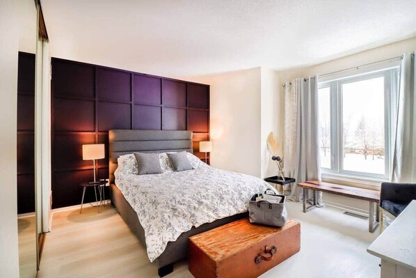 A modern bedroom with a large bed, bedside lamps, a wooden bench, and a window with curtains. - Cozy Calabogie condo with high speed internet (Calabogie)