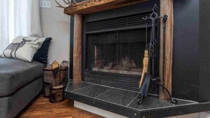 Un salon chaleureux avec une cheminée, un canapé gris et une petite table d’appoint en bois sur laquelle est posée une bougie.