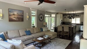A spacious living room with a large sofa, a coffee table, and a ceiling fan. There is a kitchen area with bar stools and a dining table. - Crystal Conch sleeps 16, private home close to beach access (Emerald Isle)