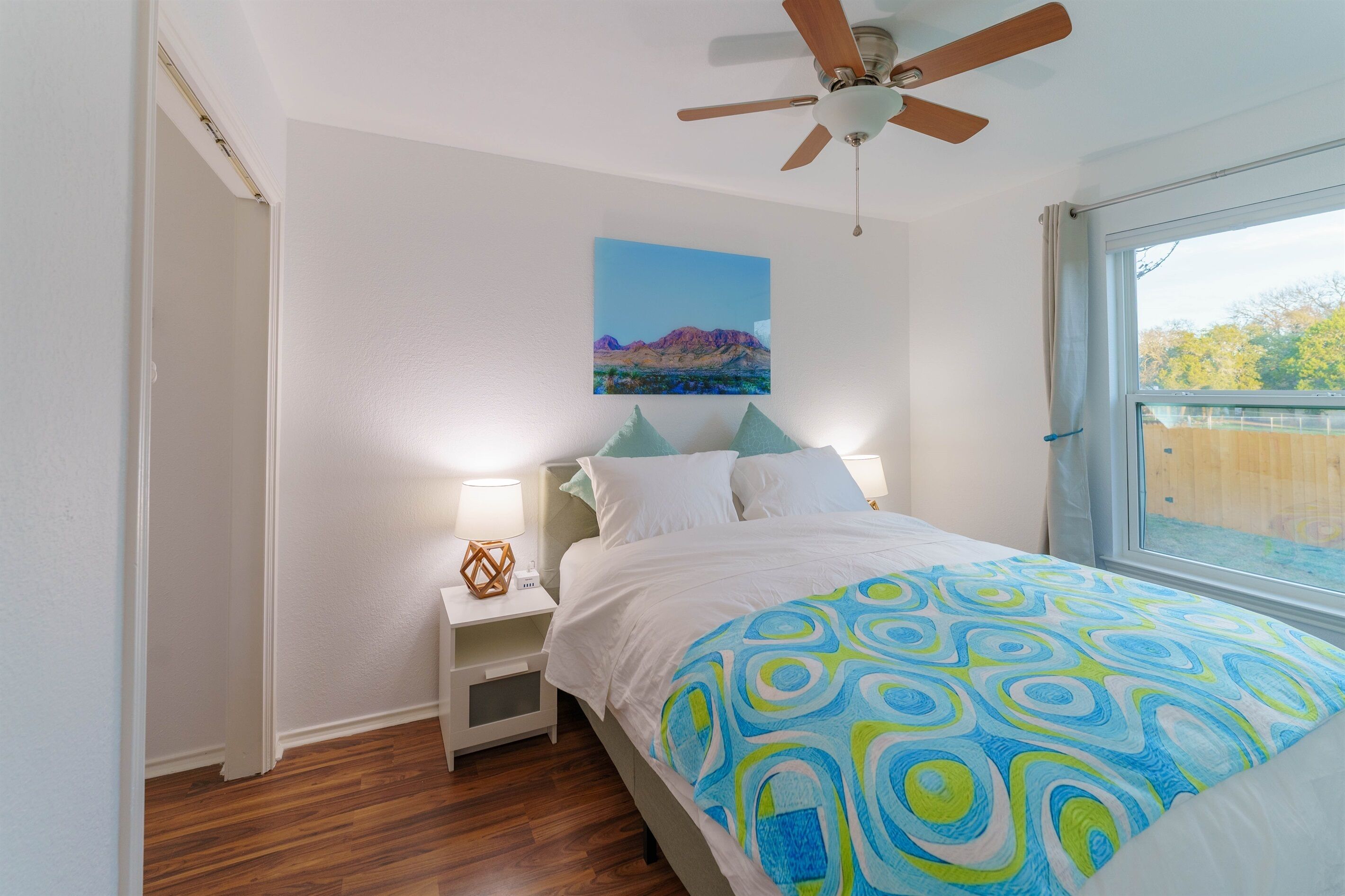 A bedroom with a bed, a nightstand, a ceiling fan, a window with a view, and a painting on the wall.