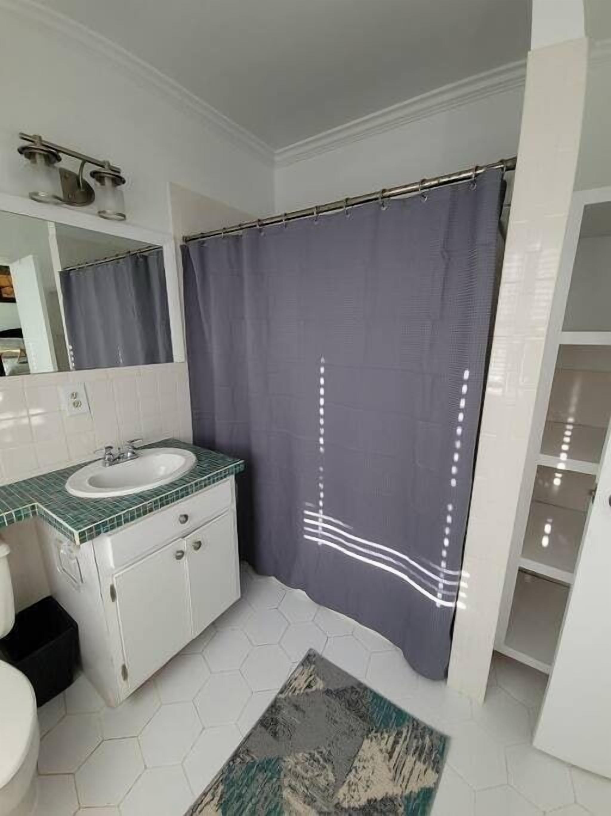 Une salle de bain avec un lavabo blanc, un plan de toilette recouvert de carreaux verts, des meubles blancs, un miroir, un rideau de douche et un sol carrelé avec des motifs hexagonaux.