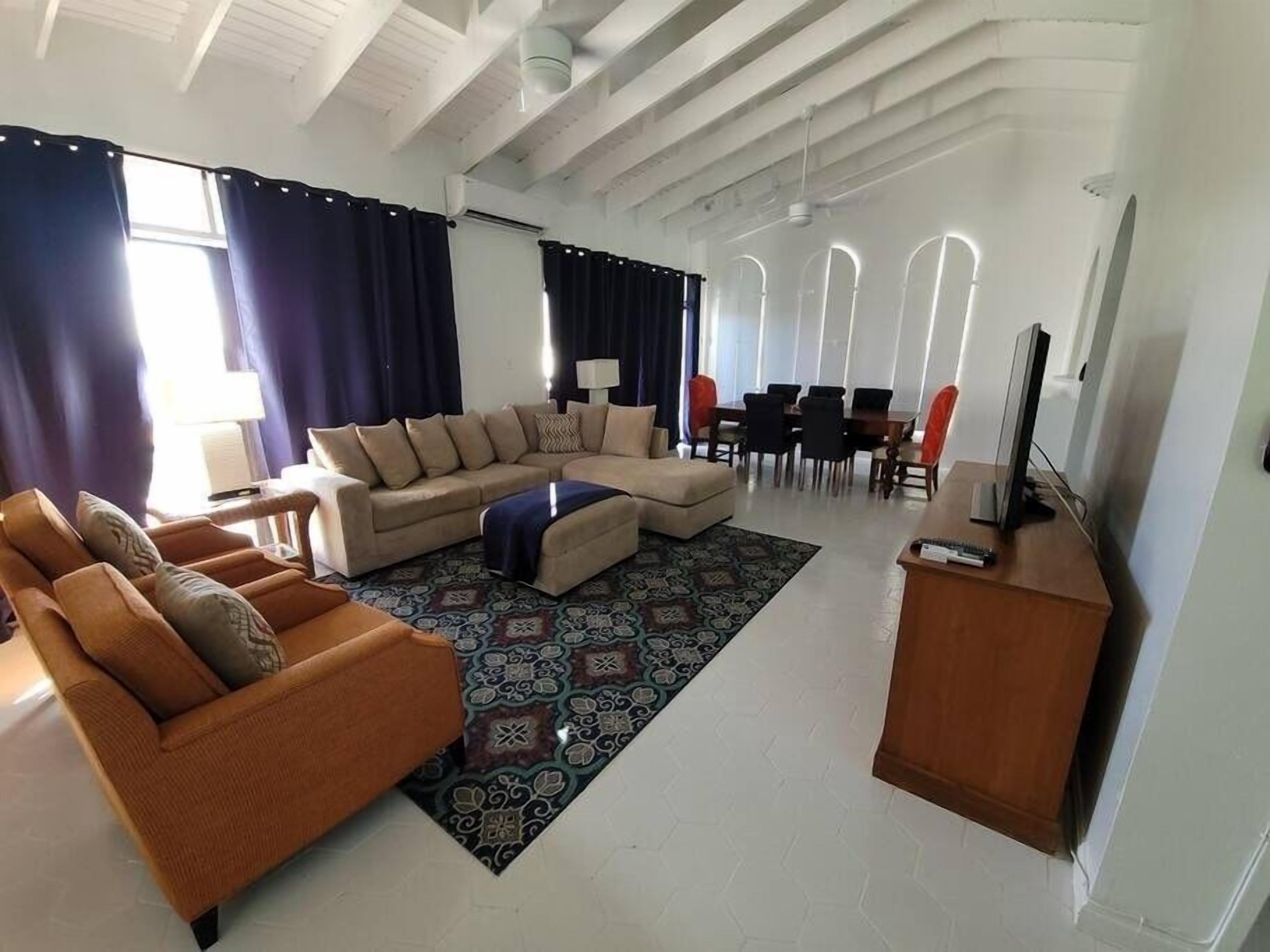 A spacious living room with a large sectional sofa, a smaller sofa, a coffee table, and a TV on a wooden stand. There is a dining area with a table and chairs in the background.