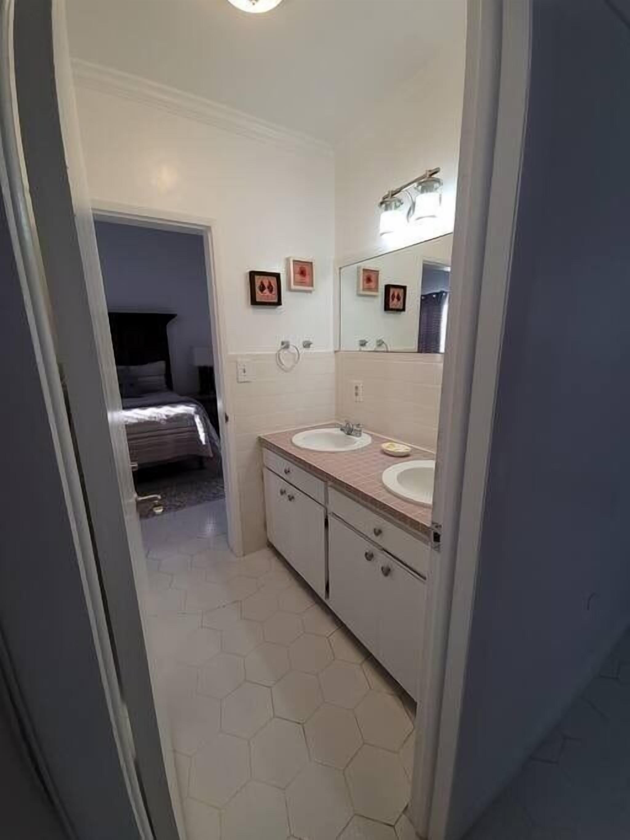 A bathroom with two sinks, a mirror, and a bed in the adjacent room.