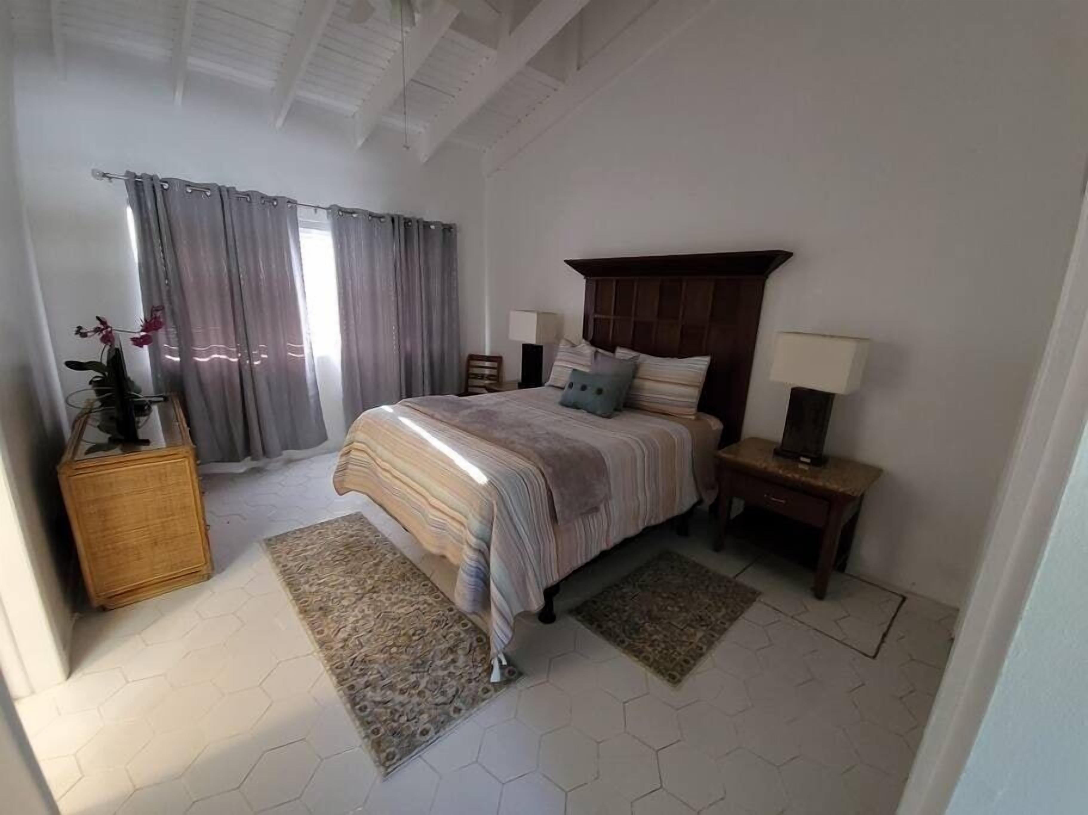 Un dormitorio con cama, mesitas de noche, cómoda, alfombra y ventana con cortinas.