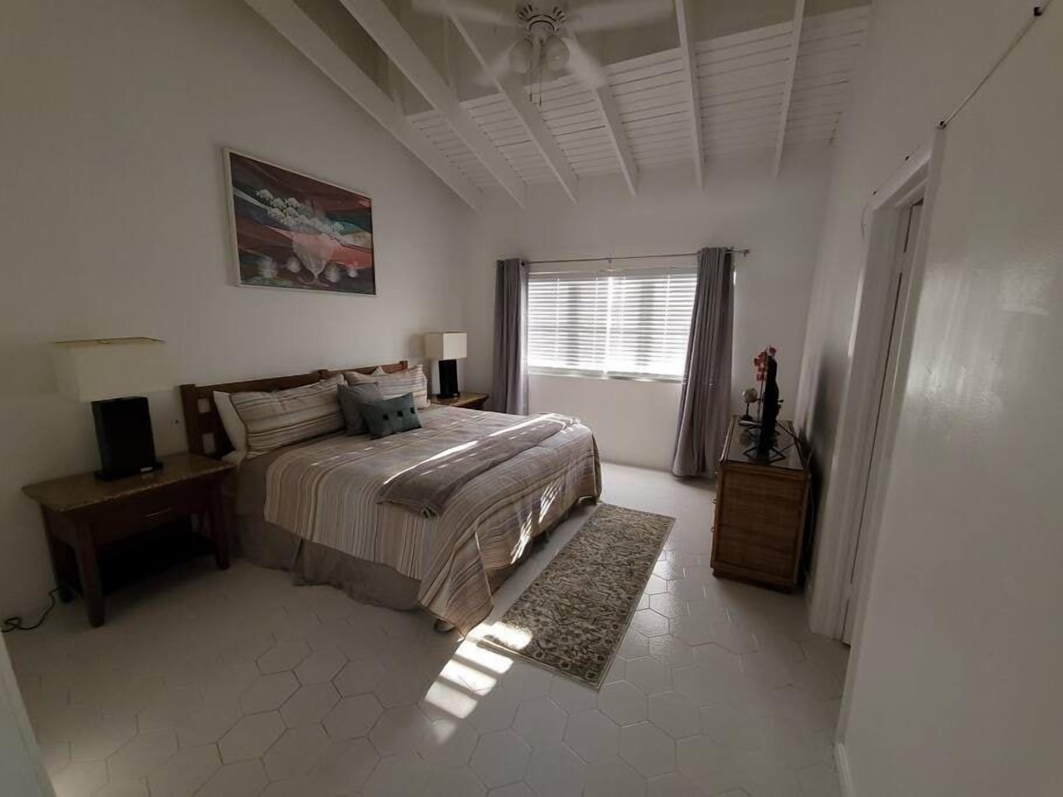 A bedroom with a bed, two bedside tables, a nightstand, a window with curtains, a ceiling fan, and a painting on the wall.