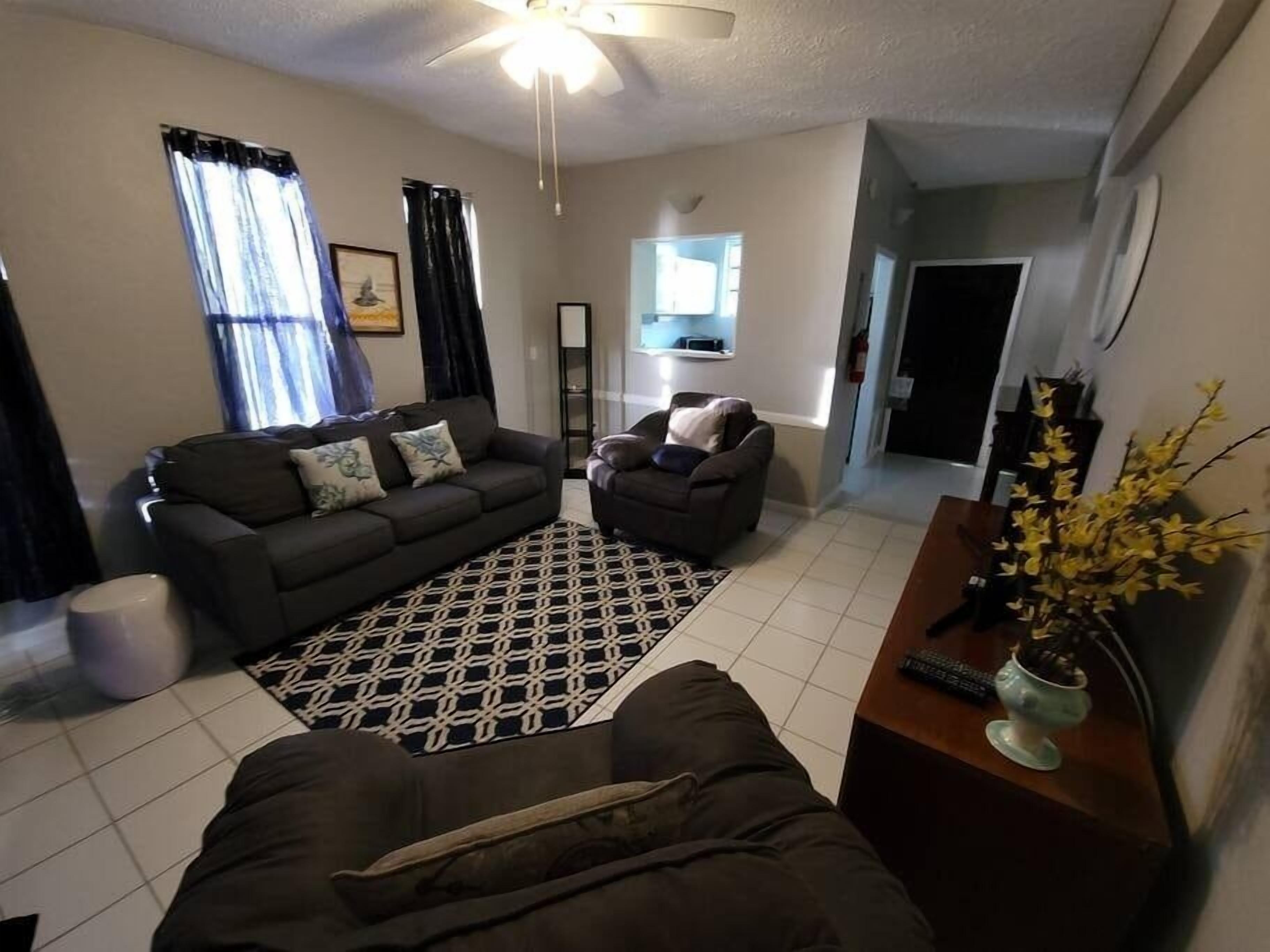 A living room with a sofa, armchair, and a coffee table. There is a ceiling fan, a window with curtains, and a plant on the side.