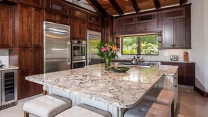 Une cuisine avec des meubles en bois foncé, des appareils électroménagers en acier inoxydable, un îlot central en granit avec des tabourets, et un ventilateur de plafond.