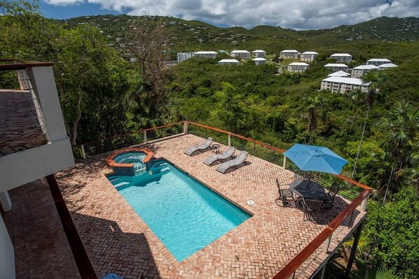 A rooftop pool with lounge chairs and an umbrella, surrounded by lush greenery and a view of distant buildings.