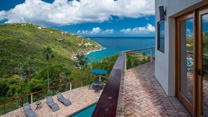 Un balcon offrant une vue sur un paysage côtier, une piscine et un espace repas aménagé avec du mobilier d’extérieur.