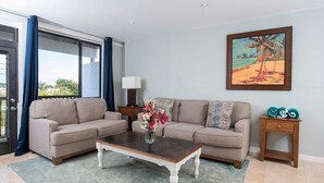 A living room with a beige sofa set, a coffee table, a painting of a beach scene, and a view through a glass door. - Blue Horizon, One - Walk to beach & Restaurants. (East End)