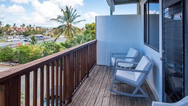 A balcony with a wooden railing, two white lounge chairs, and a view of a parking lot and palm trees. - Blue Horizon, One - Walk to beach & Restaurants. (East End)