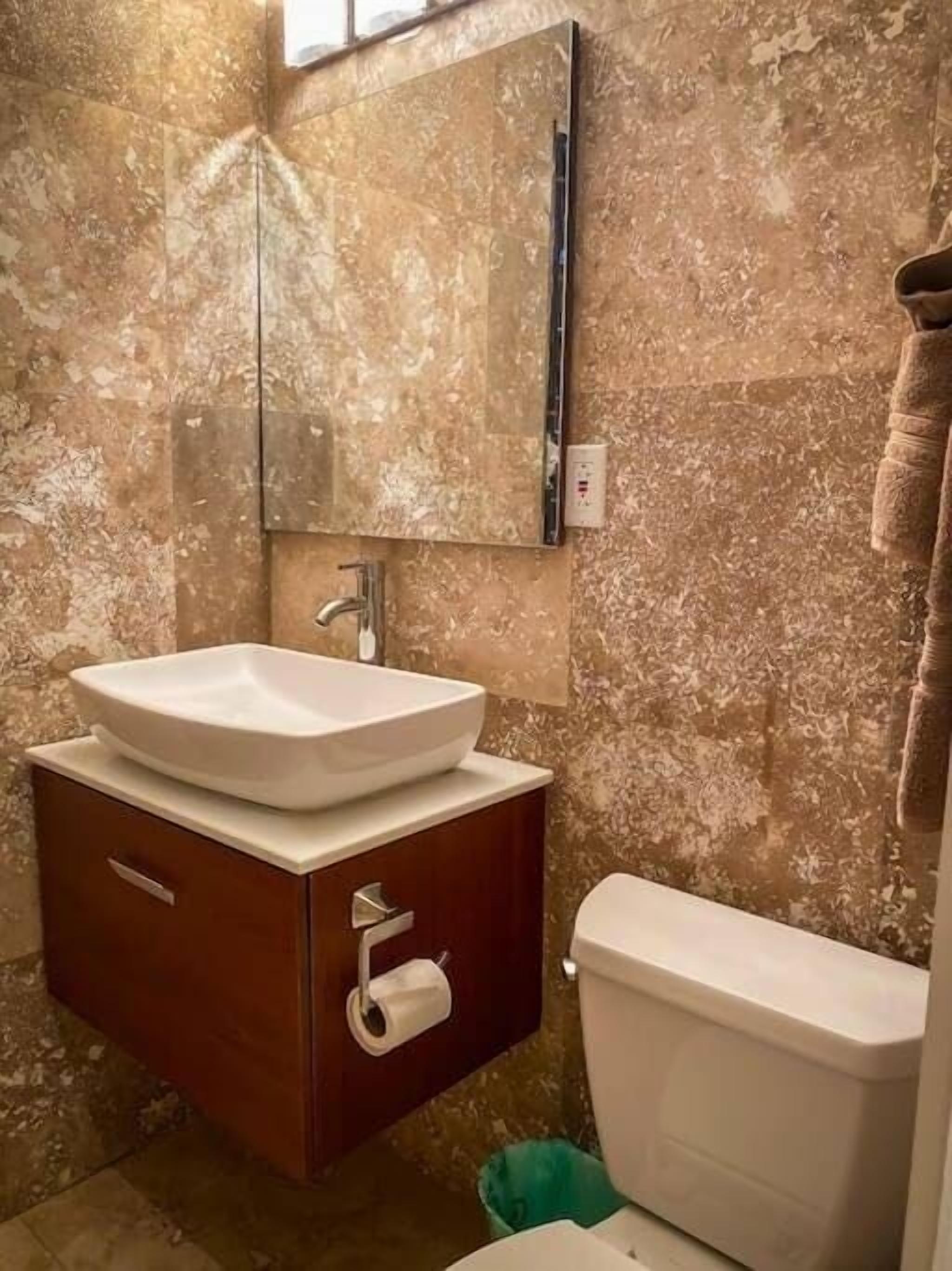 A bathroom with a marble wall, a white sink, a wooden vanity, and a toilet.