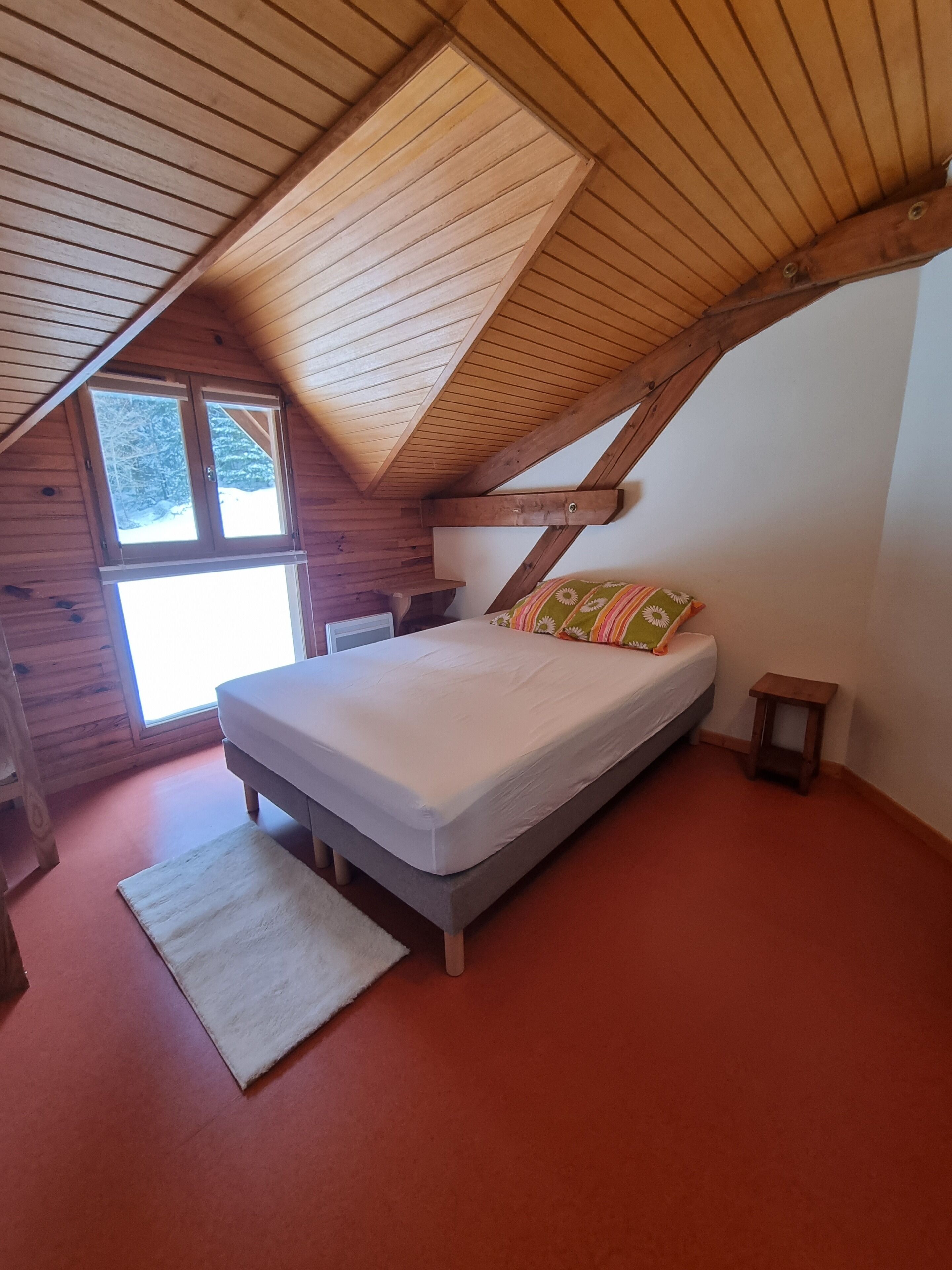Een slaapkamer met een schuine kap, een bed met een witte matras en kleurrijke kussens, een klein nachtkastje en een raam met uitzicht op bomen.