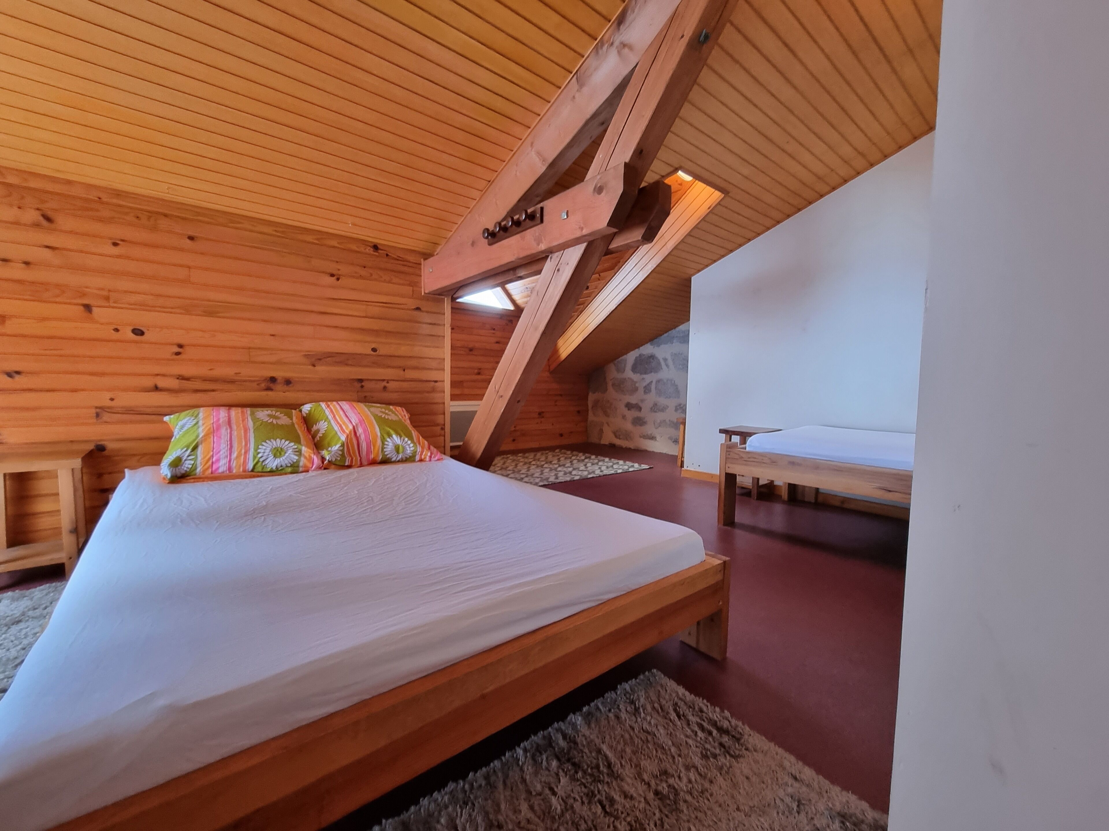 A bedroom with a wooden ceiling, a bed with colorful pillows, a stone wall, and a small table.