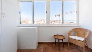 Une pièce avec une grande fenêtre, un fauteuil en osier, une petite table ronde et un radiateur blanc.