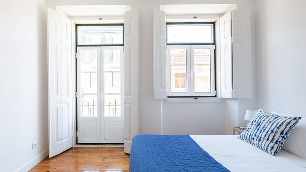 Une chambre avec un plancher en bois, un lit recouvert d’une courtepointe bleue, des murs blancs et deux fenêtres à cadres blancs.