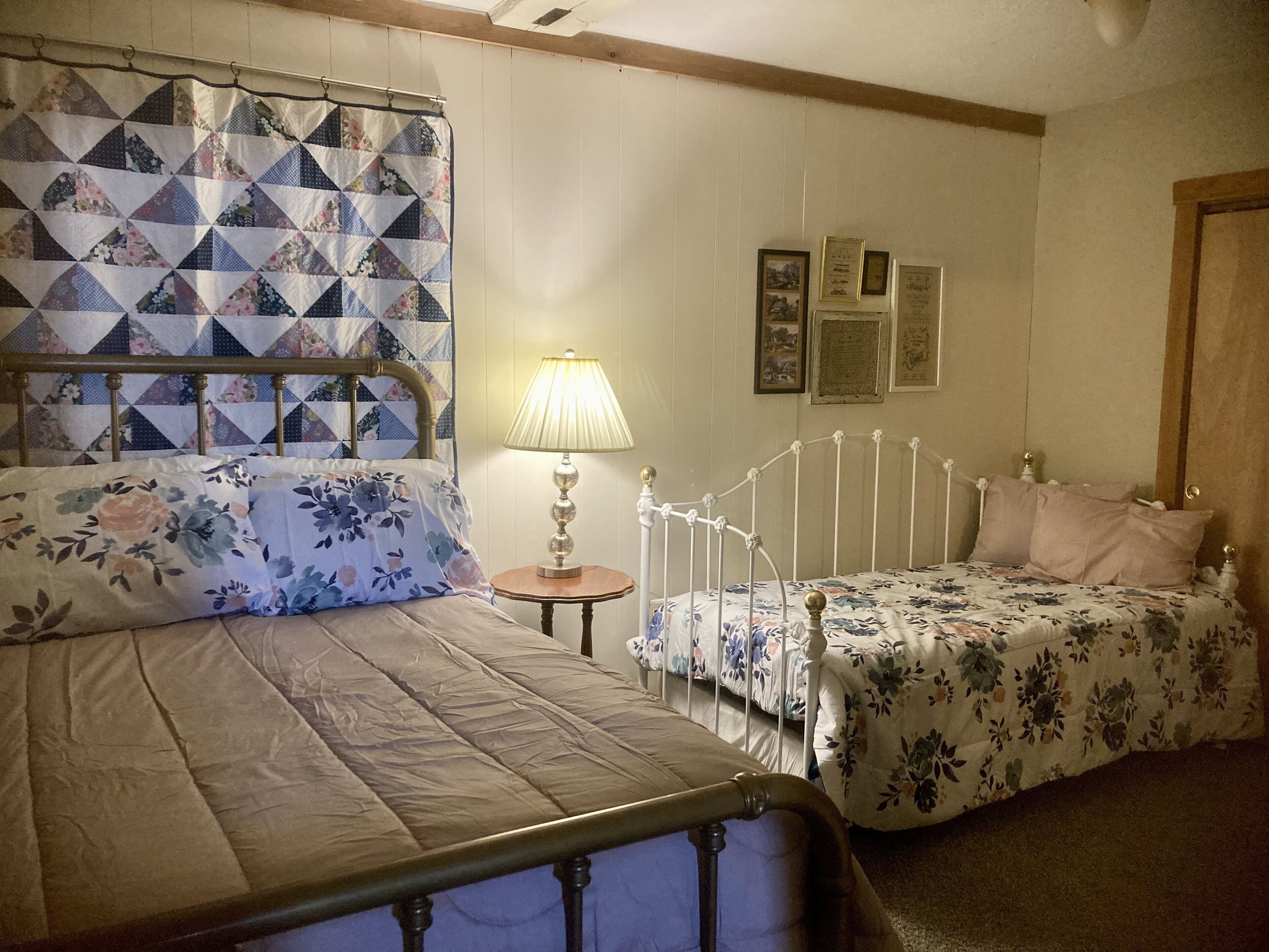 Een slaapkamer met twee bedden, een nachtkastje, een lamp en lijstjes met foto’s aan de muur.