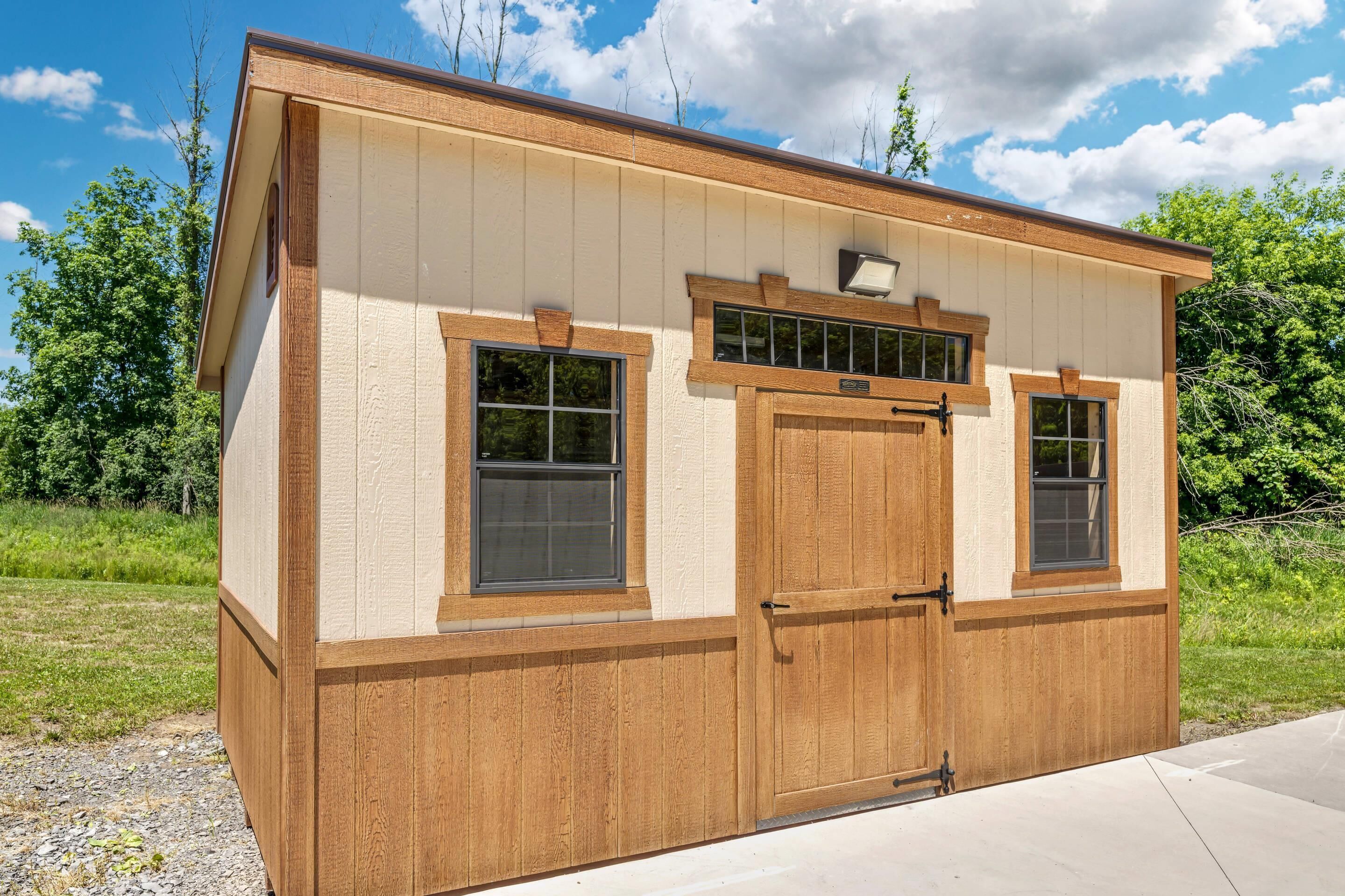 A wooden shed with two windows and a door, set in a grassy area with trees in the background.