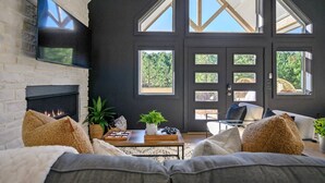 Una sala moderna con chimenea, un sofá grande y una mesa de centro. Hay varias ventanas y una puerta con paneles de vidrio.
