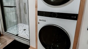 A laundry room with two stacked front-loading washing machines, a glass shower enclosure, and a black mat with "Wash Here" written on it.