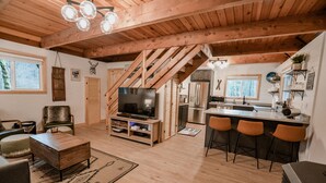 A spacious living area with a wooden ceiling, a TV on a wooden stand, a kitchen with a refrigerator, and a bar area with stools.