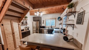 A modern kitchen with a large island, wooden ceiling beams, and a view of the outdoors through a window.